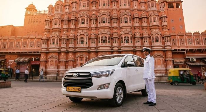 Chauffeur in white uniform standing beside a Toyota Innova Crysta outside Hawa Mahal in Jaipur